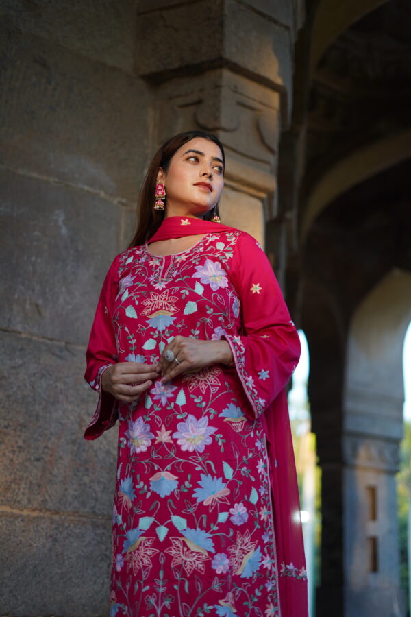 Woman in a magenta Aari-embroidered kurta, leaning gracefully against a stone pillar.