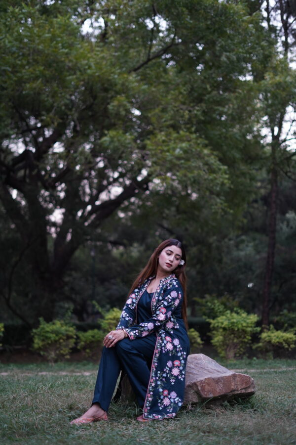 Woman seated in a park wearing a dark teal three-piece jacket set with floral embroidery.