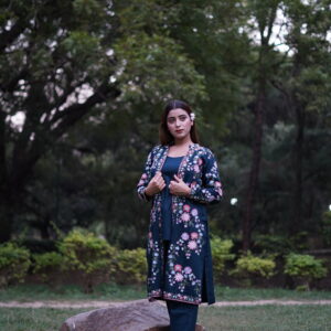 Woman wearing a dark teal Aari-embroidered jacket set, standing in a serene park setting.