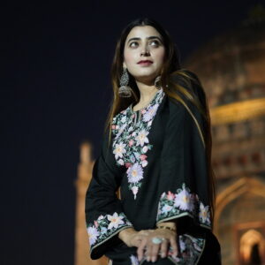 Woman wearing a black short kurta with colorful Aari embroidery, sitting gracefully against a night backdrop.